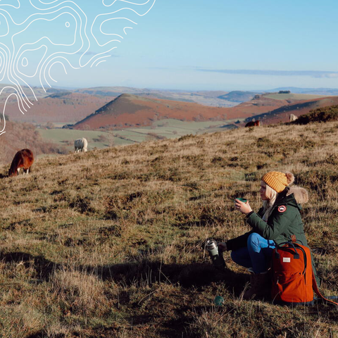 Woman sat on hilltop with grazing ponies in the distance