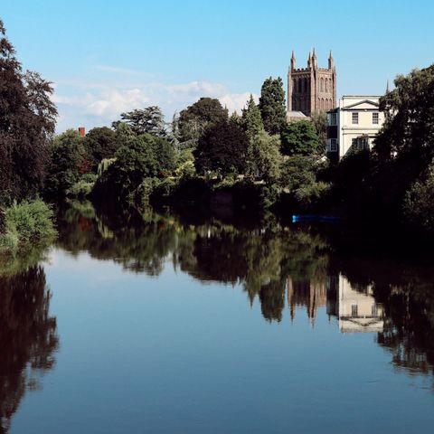 A view across a very still River Wye towards the Cathedral with the building, blue skies and trees reflected in the water
